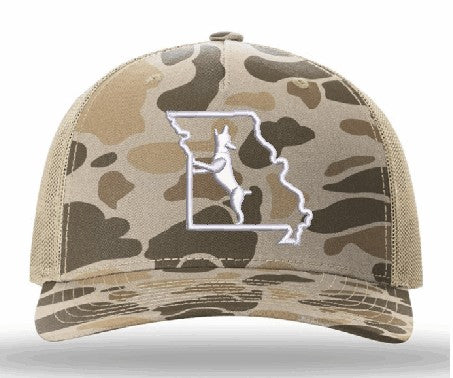 A camouflage trucker hat with a tan and brown camo pattern, featuring a white embroidered outline of Missouri with a white dog silhouette on the front panel.