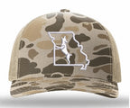 A camouflage trucker hat with a tan and brown camo pattern, featuring a white embroidered outline of Missouri with a white dog silhouette on the front panel.